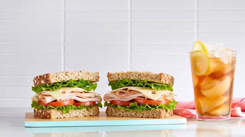 A homemade sandwich sliced in half on a cutting board, next to an iced beverage with lemon in a tall glass.
