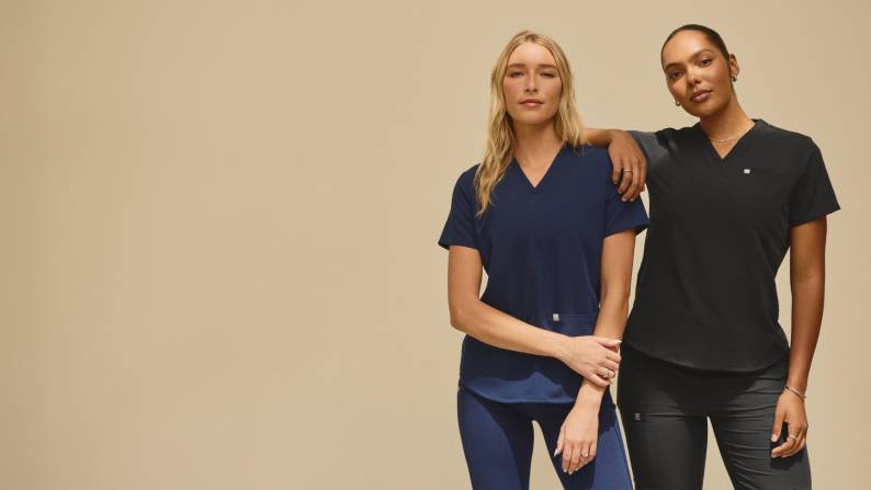 A woman wearing a navy blue set of scrubs and a woman wearing a black set of scrubs are leaning against one another.
