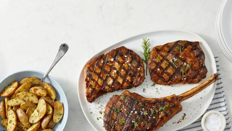 Grilled steaks on a white plate with roasted potato wedges, herbs and a small dish on a light surface.