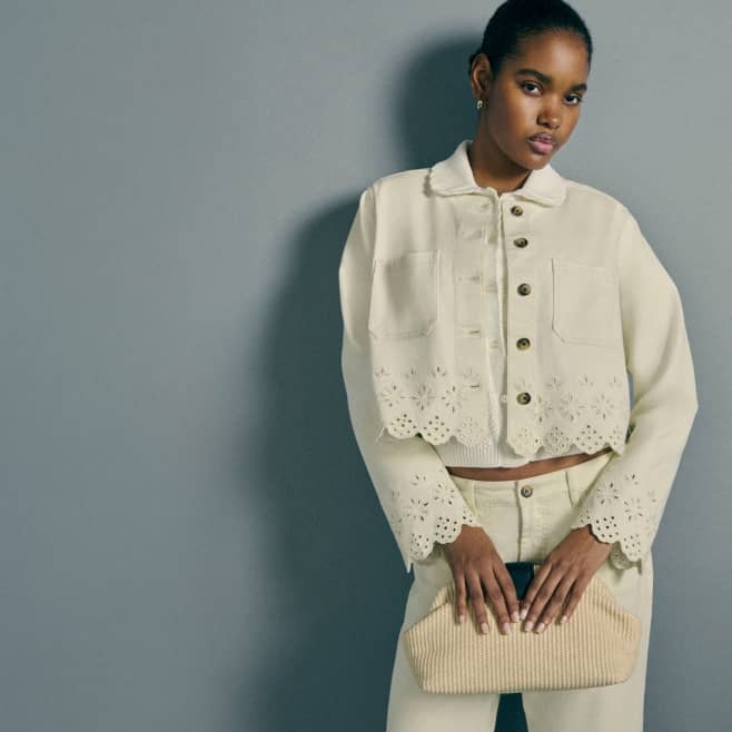 A woman wears a cream eyelet jacket and matching denim pants, holding a woven clutch.