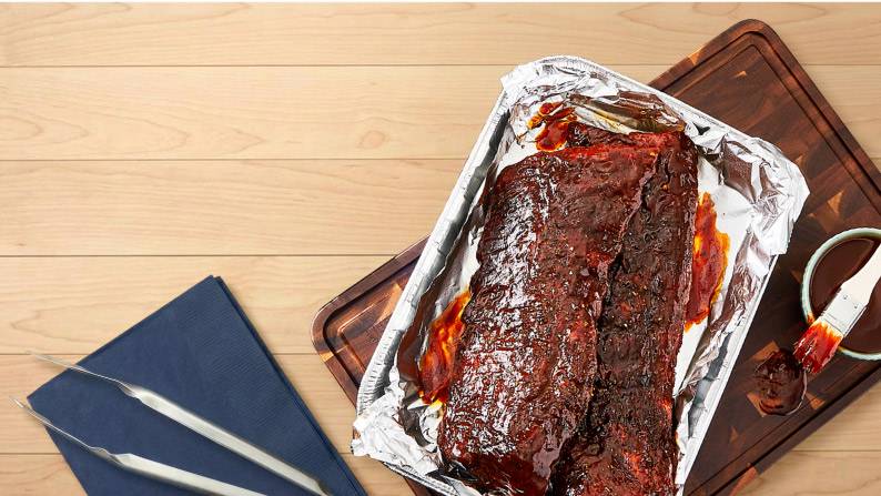 Barbecue ribs coated in sauce on foil atop a tray with a basting brush, small bowl of sauce and napkin on a wood surface.