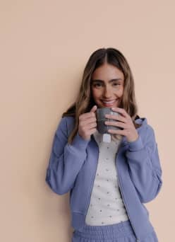 Adult in light purple loungewear holds a mug of tea and smiles.