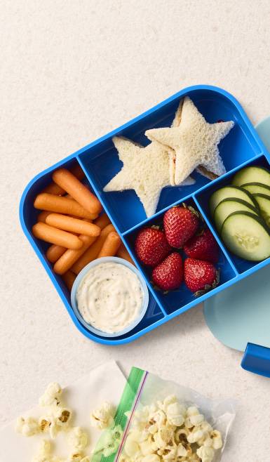 A bag of popcorn next to a Bentgo box of baby carrots, dip, strawberries, star-shaped sandwiches, and cucumber slices.