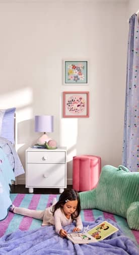 A girl reads on a striped rug. A white nightstand with a purple lamp, pink ottoman and green backrest pillow are styled in a brightly lit bedroom.