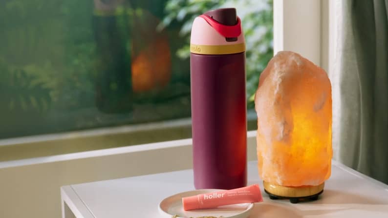 A maroon insulated water bottle stands next to a glowing pink Himalayan salt lamp and a small tube of lip butter on a white table.