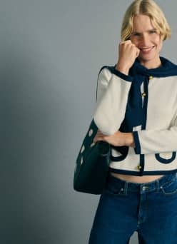 A smiling fashion model wears a cream cardigan with navy trim, blue jeans and carries a black bag against a gray background.