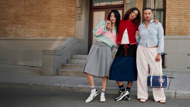 AÂ woman in a rugby shirt and a gray skirt, a woman in a red cardigan and a white shirt, and another wears a blue dress shirt and holds a blue bag.