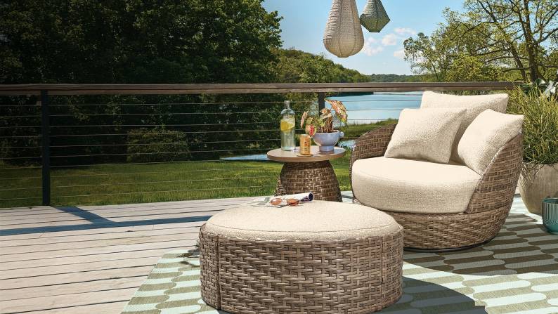 A woven wicker swivel chair and ottoman with cream cushions are on a patio overlooking a lake and trees.