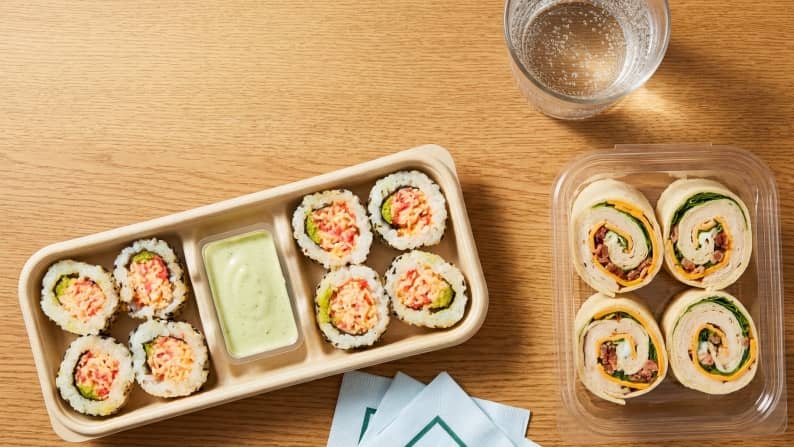 Assorted sushi rolls and wraps in clear containers on a wooden surface.