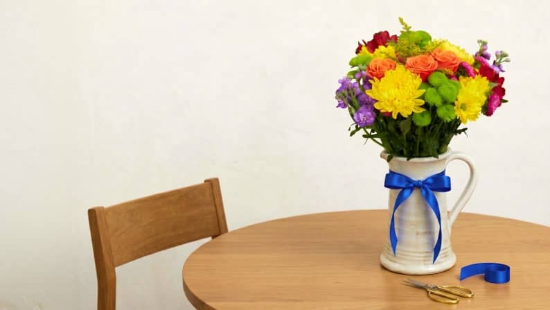 A bouquet of purple, red, pink, yellow, orange, and green flowers in a white pitcher vase with a blue ribbon tied around it.