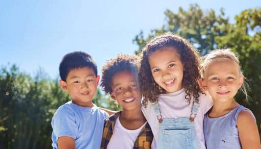 Four smiling children stand close together outdoors in casual summer clothing.
