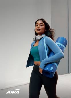 A young woman is shown wearing an Avia sports bra, blue zip-up jacket and black leggings. She is carrying a yoga mat.