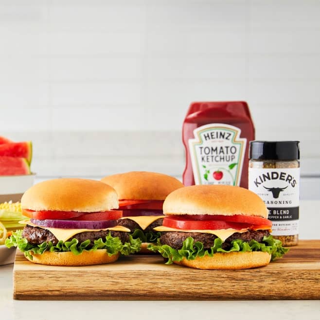 Cheeseburgers with lettuce, tomato, and onion next to Heinz ketchup and Kinder’s seasoning. Sliced watermelon is in the background.