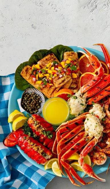 A platter of grilled salmon topped with fruit salsa, crab legs, lobster tails, shrimp, cut citrus fruit, capers, and melted butter.