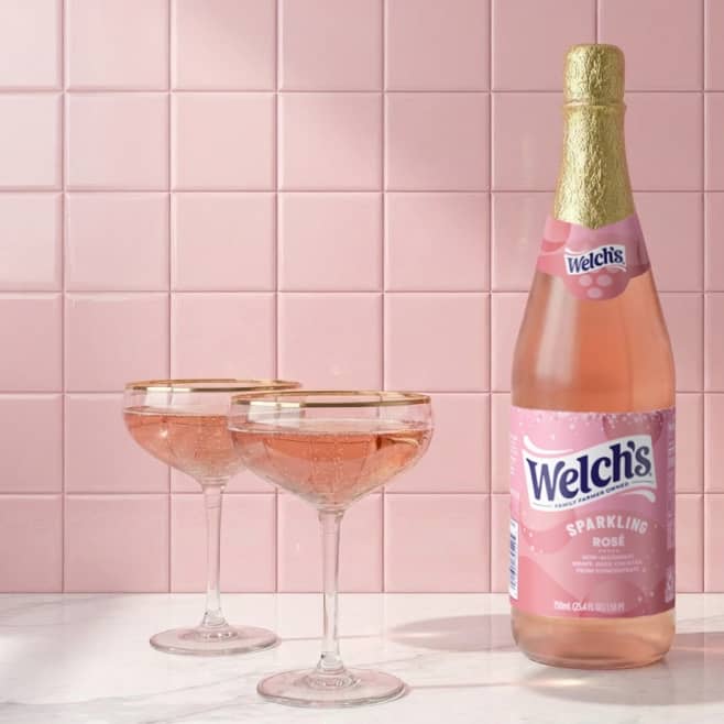 A bottle of Welch’s sparkling rose beverage sits on a kitchen counter next to two coupe glasses filled with a pink fizzy drink.