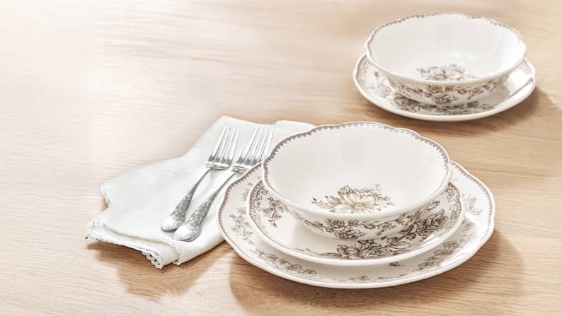 Cream-colored dinnerware with a grey floral print are shown on a wood tabletop. Also shown is a cloth napkin, silverware and a vase with flowers.