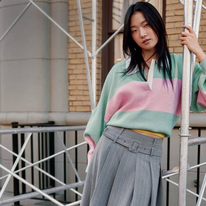 Model wearing a pastel rugby-style top and high-waisted pleated trousers, leaning against scaffolding in an urban setting.