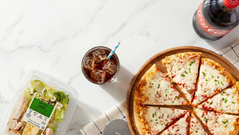 A pizza, a Marketside Caesar salad, a bottle of Coca Cola, and a glass with ice and a straw.