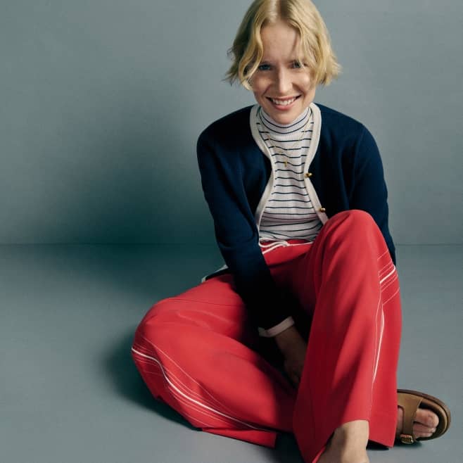 A smiling fashion model sits on the floor wearing a striped top, navy cardigan, red trousers and brown sandals against a gray background.