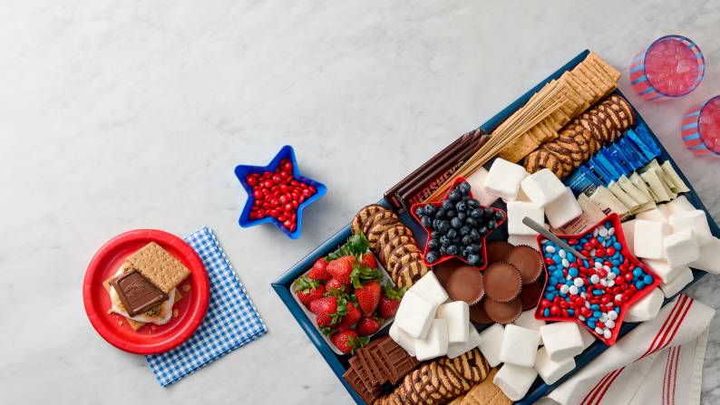 A s’more on a paper plate next to a tray of berries, chocolate bars, marshmallows, cookies, candies, graham crackers, and skewers.