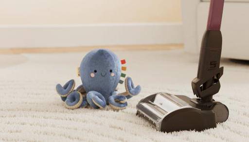 Blue plush octopus toy sits on a light carpet beside a vacuum cleaner head.