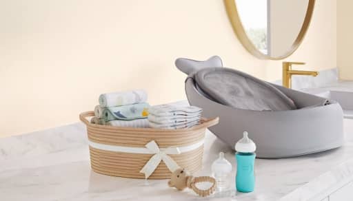 Baby changing station with basket of diapers and blankets beside bottles, pacifier and teether on a marble counter.