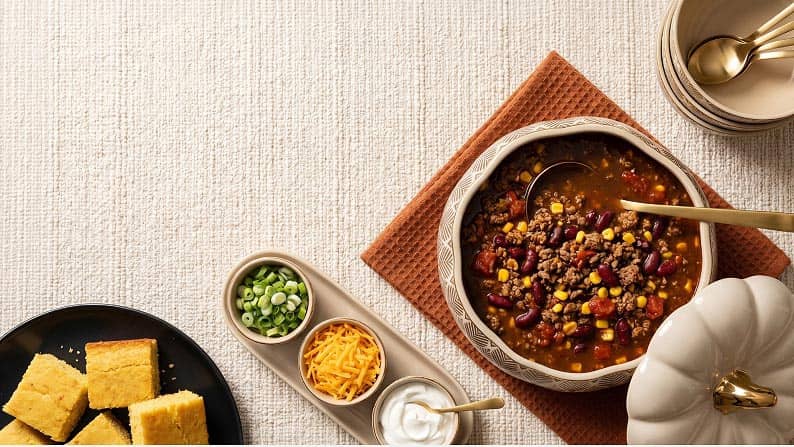 Chili in a pumpkin-shaped serving bowl next to bowls of green onions, cheddar cheese, sour cream, and a plate of cornbread on an off-white tablecloth.