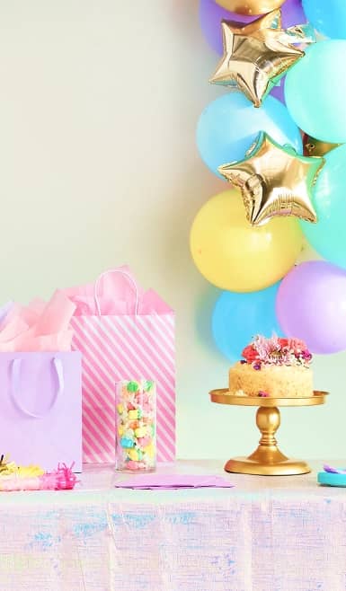Party scene with blue and gold balloons alongside pink gift bags and a cake on a stand.