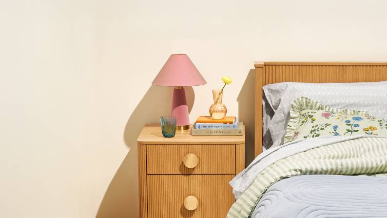 A pink lamp, blue glass and yellow flower on a fluted wood nightstand next to a bed with floral and striped linens and a blue bedspread.