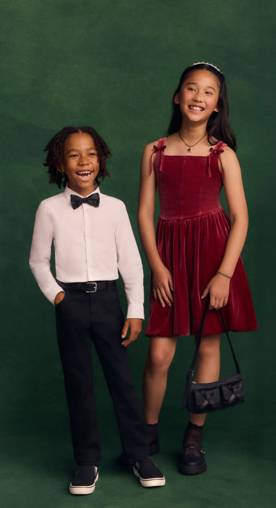 Two children wearing festive holiday outfits are pictured on a green backdrop.