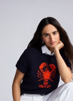 A woman wears a short-sleeved sweater with a red lobster graphic on it, a white-collar shirt underneath, and white pants.
