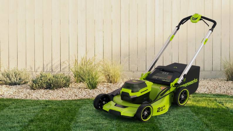 A lawnmower on green grass, in front of a garden and fence.