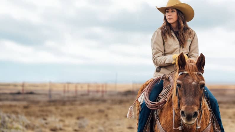 A female rancher wearing a large hat rides a horse.