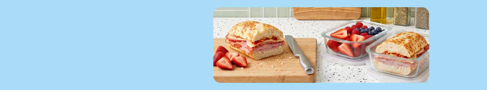 A cutting board with strawberries and half a sub sandwich next to glass containers of strawberries and blueberries and the remaining half sub.