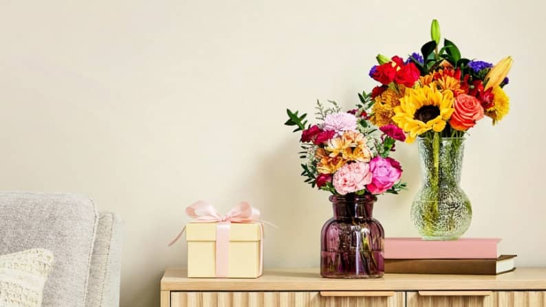 A yellow gift box with a pink ribbon next to two vases full of colorful flowers and greens on a side table.
