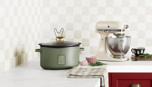 An electric stand mixer is on a kitchen countertop next to a sage green slow cooker and a checkered dish towel.