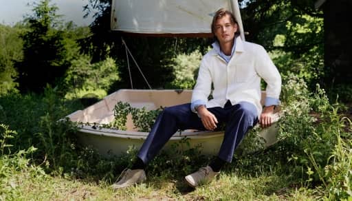 Man wearing a white overshirt over a light blue button-up and navy pants, sitting on a sailboat.
