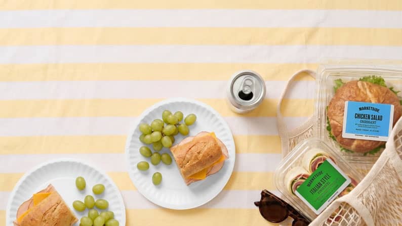 Two paper plates with half subs and green grapes next to a Marketside chicken salad croissant and Marketside Italian style pinwheels.