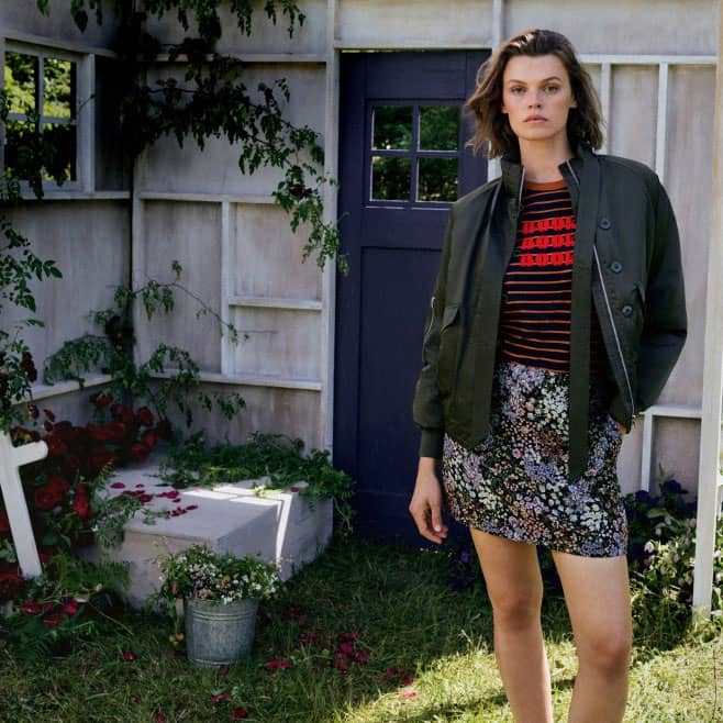 Woman in a dark green jacket, striped top and floral jacquard mini skirt, standing in front of a house.
