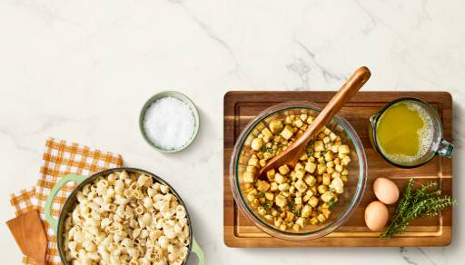 A bowl of macaroni and cheese is shown next to a bowl of stuffing on a cutting board with eggs, herbs, a cup of broth, and a small bowl of salt.