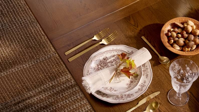 Gold-tone flatware, floral dinnerware, glassware, and a rolled-up napkin are set on a dark wooden dining table.