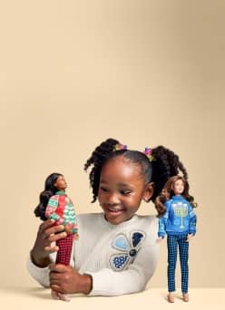 Smiling child in a sweater holds a fashion doll while another doll stands on the table.
