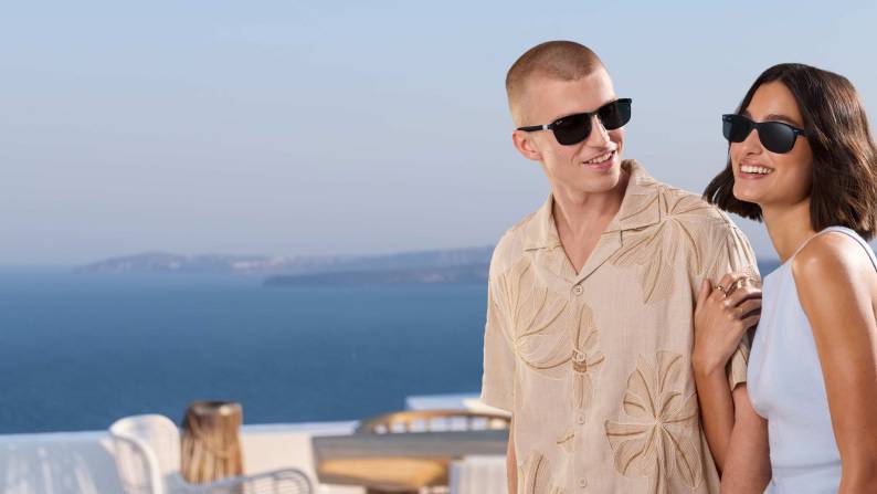 Two people wearing black sunglasses standing outdoors near the ocean.