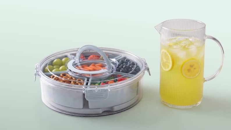 A divided snack container filled with fruit, pretzels and candy beside a ribbed glass pitcher. From nine dollars and eighty-seven cents.