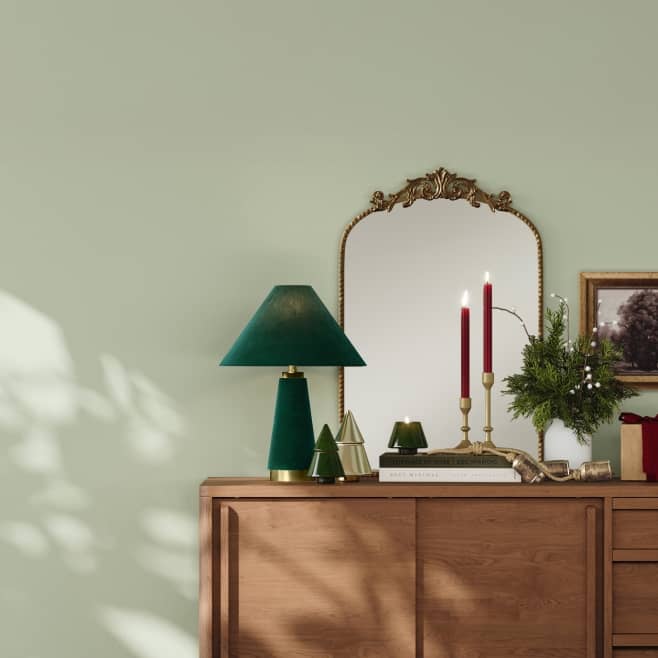 A wooden console is shown with a large mirror, a green lamp and various decor against a sage wall.