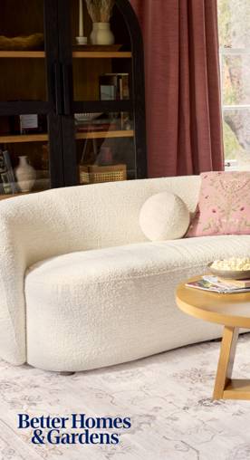 A white curved upholstered couch is seen with a wooden coffee table. A glass-enclosed cabinet sits next to a window with red curtains.