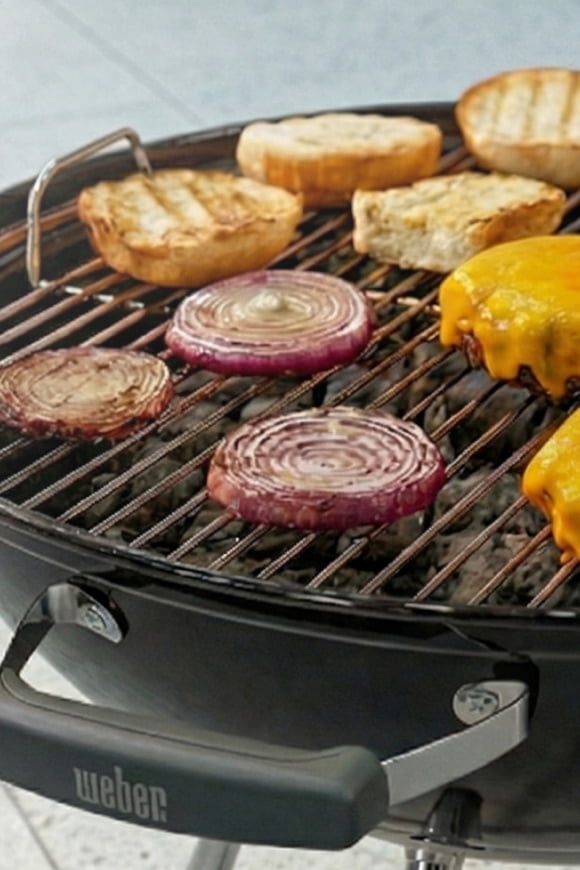 View cheeseburgers, onion slices and buns cooking on a Weber charcoal grill.