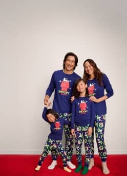 A family of four wearing matching Snoopy pajamas is pictured on a beige backdrop.