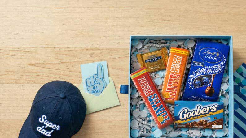 Gift box with assorted candies and chocolates next to a Super dad baseball cap and a Number 1 Dad foam finger on a wood surface.