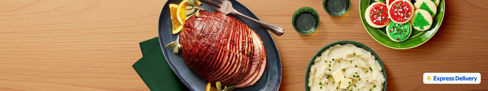 Sliced ham on a platter with herbs and corn, next to a plate of iced and tree-shaped cookies. An Express Delivery badge is shown.